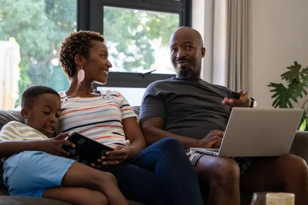 A smiling family of three sits on a dark sofa in a bright living room, with the father using a laptop, the mother looking toward him, and their young son playing on a tablet.